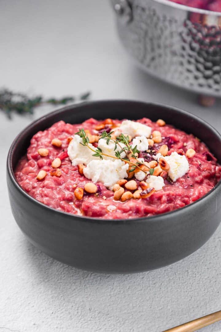 A black bowl filled with creamy beet risotto, topped with crumbled cheese, pine nuts, and a sprig of thyme, sits on a light gray surface with a metal pot blurred in the background.