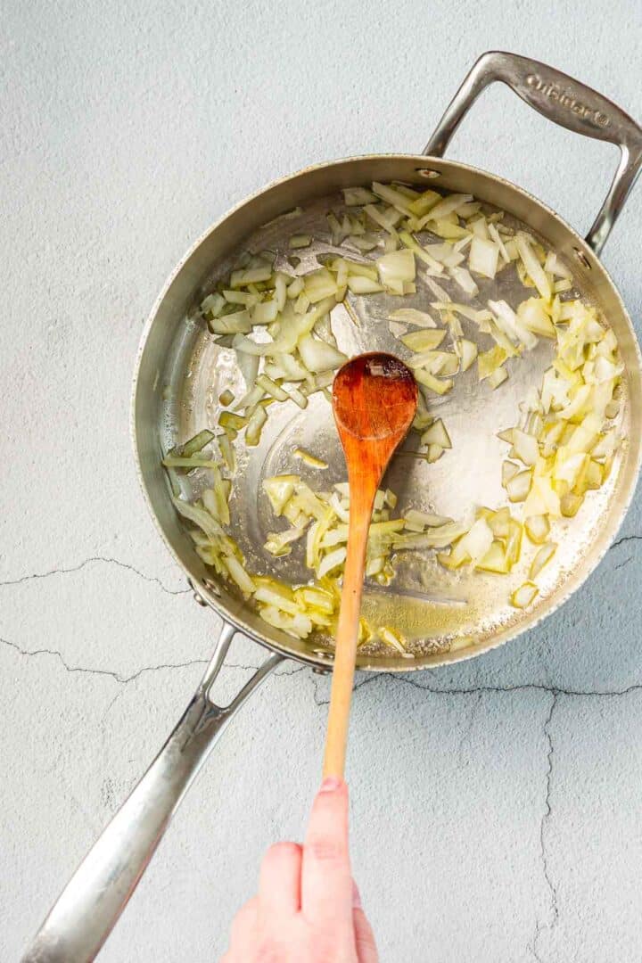 A hand stirs chopped onions in a stainless steel pan with a wooden spoon. The onions are being sautéed in oil on a light gray, textured surface with visible cracks.
