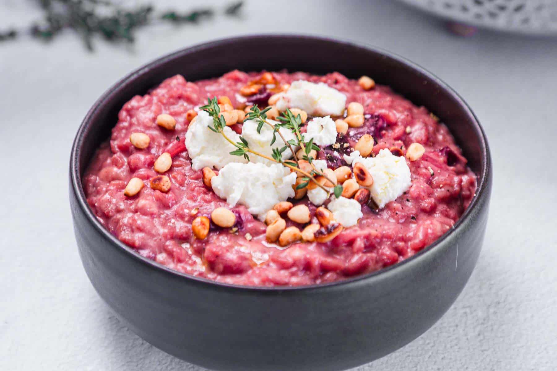 A black bowl filled with vibrant beetroot risotto, topped with crumbled white cheese, toasted pine nuts, and fresh thyme sprigs, placed on a light surface.