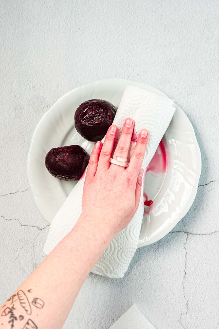 A hand holding a paper towel is blotting two peeled beets on a white plate. The paper towel is stained with beet juice, and part of a tattooed arm is visible in the lower left corner.