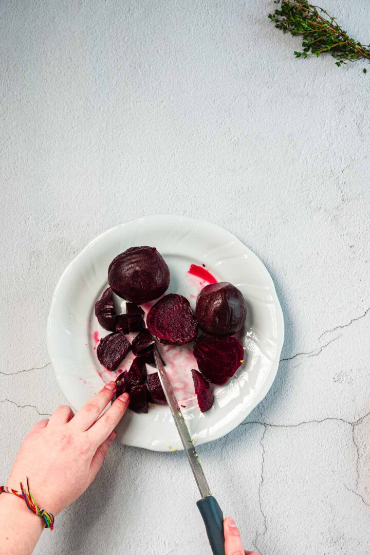 A hand uses a knife to slice cooked beets on a white plate. The plate is placed on a light gray surface, with a small bunch of herbs visible in the top right corner.