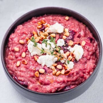 A bowl of creamy pink Beetroot Risotto topped with crumbled cheese, toasted pine nuts, and fresh thyme sprigs sits on a light surface.