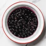 A white bowl with a red rim contains homemade blueberry sauce with stewed blueberries in purple liquid, placed on a white marble surface.