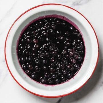 A white bowl with a red rim contains homemade blueberry sauce with stewed blueberries in purple liquid, placed on a white marble surface.