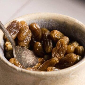 A ceramic bowl filled with golden raisins, with a metal spoon resting inside.