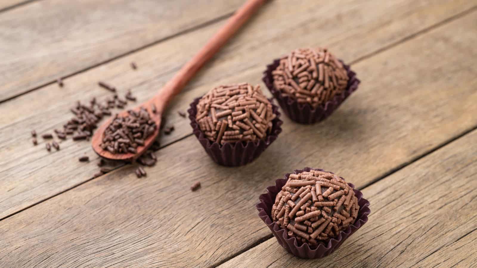 Three chocolate brigadeiros with chocolate sprinkles sit on a wooden surface. A wooden spoon with chocolate sprinkles rests nearby, with some sprinkles scattered on the table.