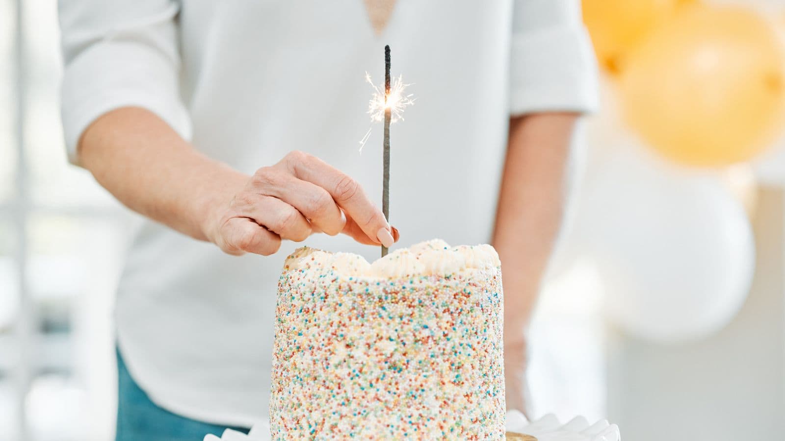 A person holds a lit sparkler on top of a cake covered in colorful sprinkles. Yellow and white balloons are visible in the blurred background.