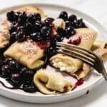 A plate of cheese blintzes filled with creamy cheese, topped with blueberry compote and powdered sugar. A fork is cutting into one of the blintzes, revealing the luscious filling inside.