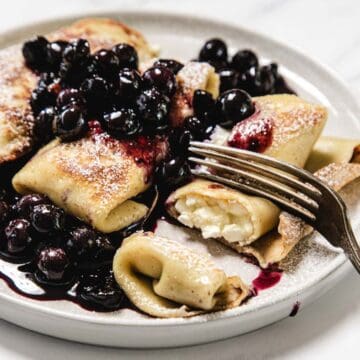 A plate of cheese blintzes filled with creamy cheese, topped with blueberry compote and powdered sugar. A fork is cutting into one of the blintzes, revealing the luscious filling inside.