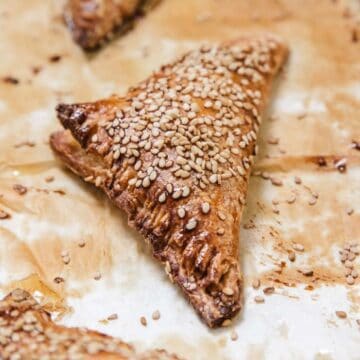 A baked triangular Cheese Bourekas pastry topped with sesame seeds sits on a sheet of parchment paper, with other similar pastries partially visible in the background.