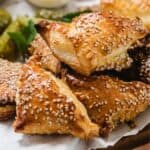 A close-up of several golden-brown Cheese Bourekas triangles topped with sesame seeds, stacked on parchment paper, with part of a pickle and mint leaves visible in the background.