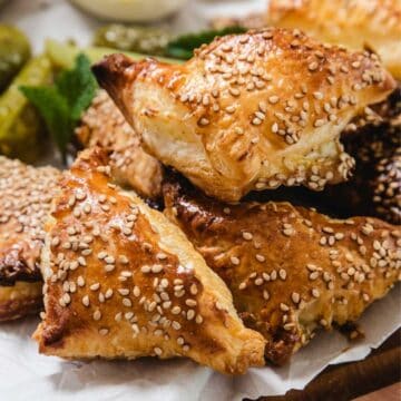 A close-up of several golden-brown Cheese Bourekas triangles topped with sesame seeds, stacked on parchment paper, with part of a pickle and mint leaves visible in the background.