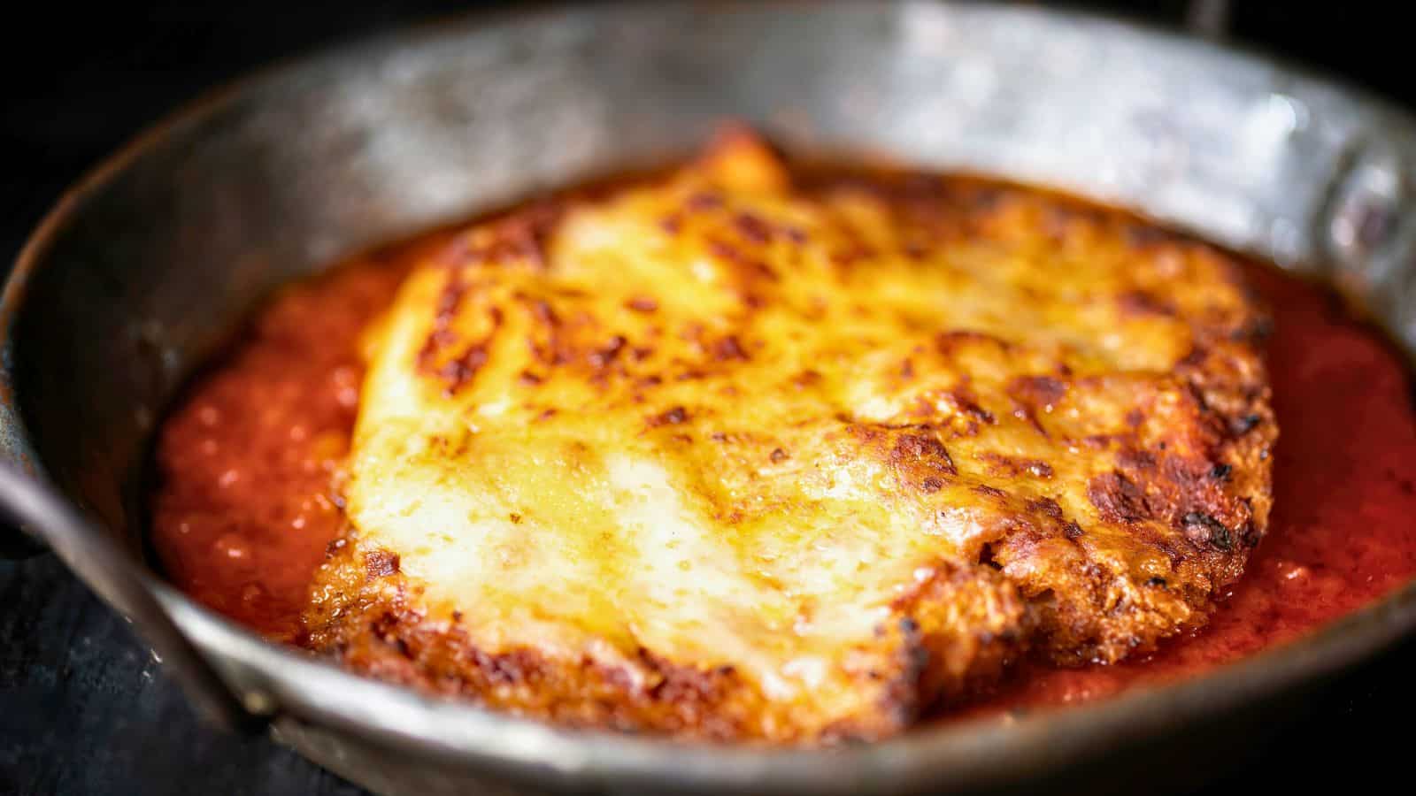 A metal pan holds a breaded cutlet topped with melted, browned cheese, sitting in a pool of red tomato sauce.