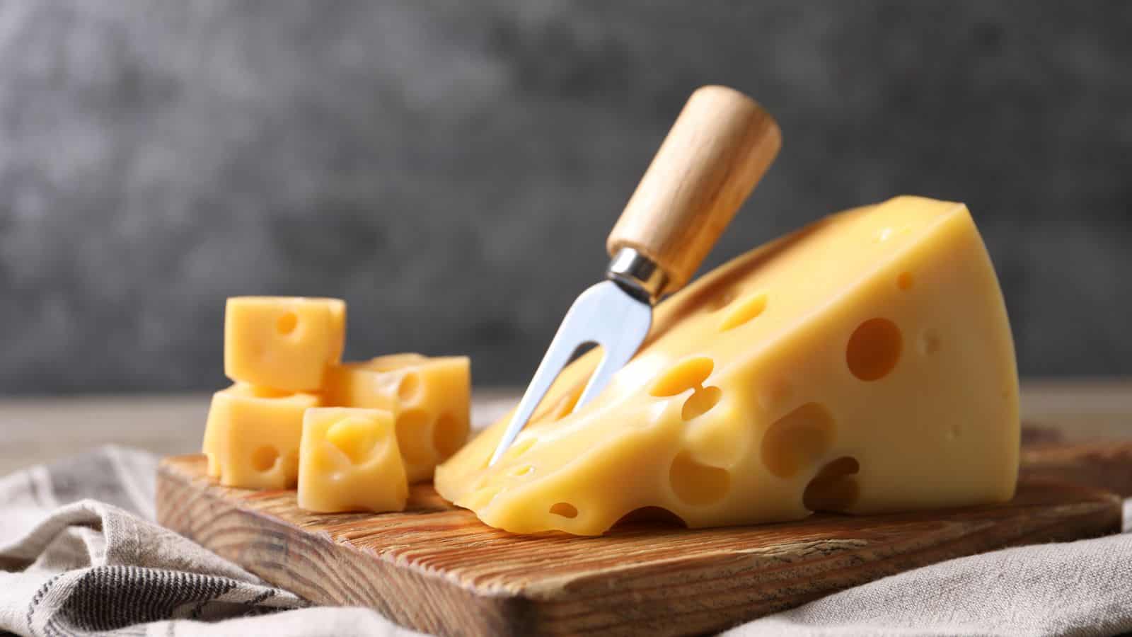 A wedge of Swiss cheese with characteristic holes sits on a wooden board, accompanied by small cubes of cheese and a cheese fork with a wooden handle. A beige cloth is partially visible underneath the board.