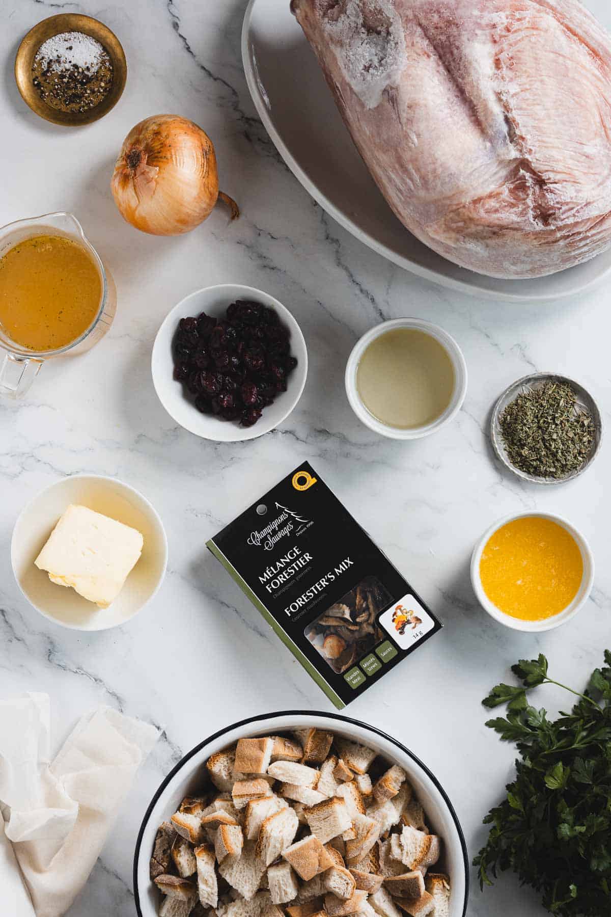 Overhead view of various ingredients on a marble surface, including a raw turkey wrapped in cheesecloth, chopped bread, butter, onion, dried cranberries, broth, melted butter, white wine, dried herbs, a mushroom mix packet, and fresh parsley.