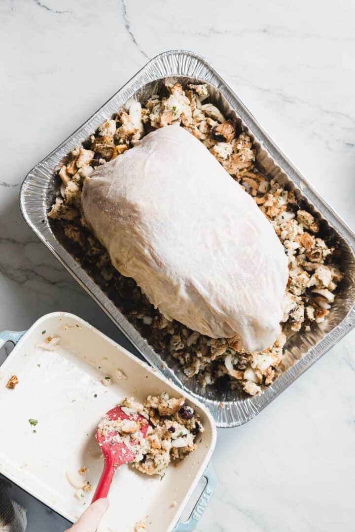 A raw turkey breast rests on a bed of gluten free stuffing in an aluminum roasting pan. Next to it, a hand uses a red spatula to scoop more gluten free stuffing from a ceramic dish onto the pan, all set on a white countertop.
