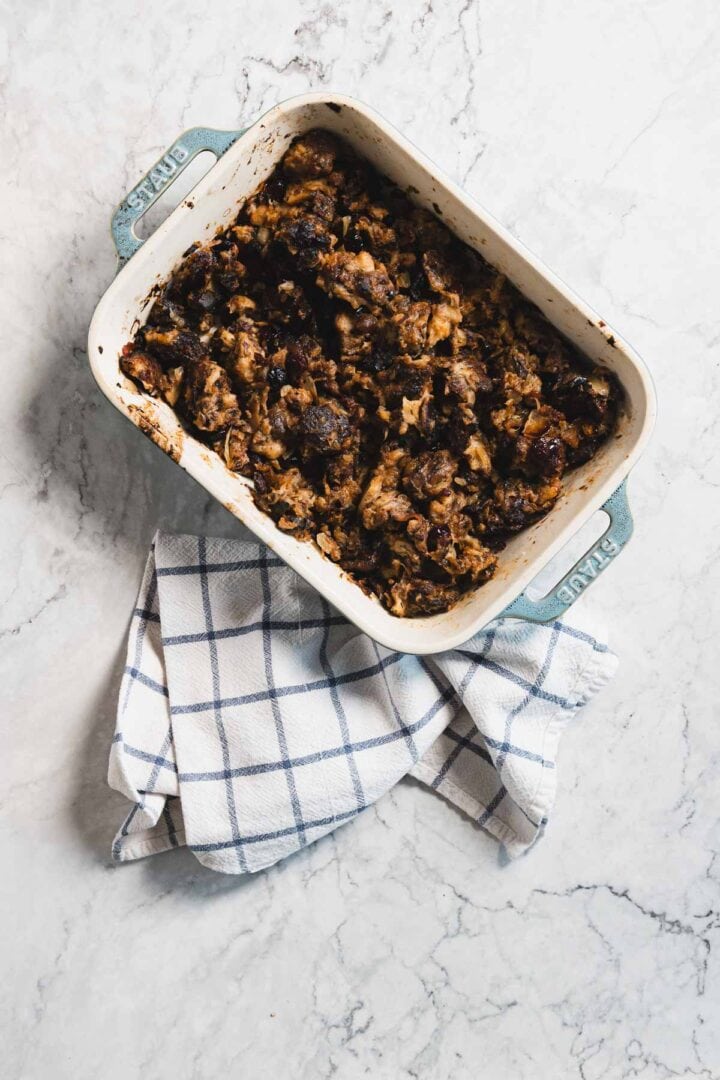 A rectangular baking dish filled with dark, baked gluten free stuffing sits on a folded checkered kitchen towel atop a marble countertop.