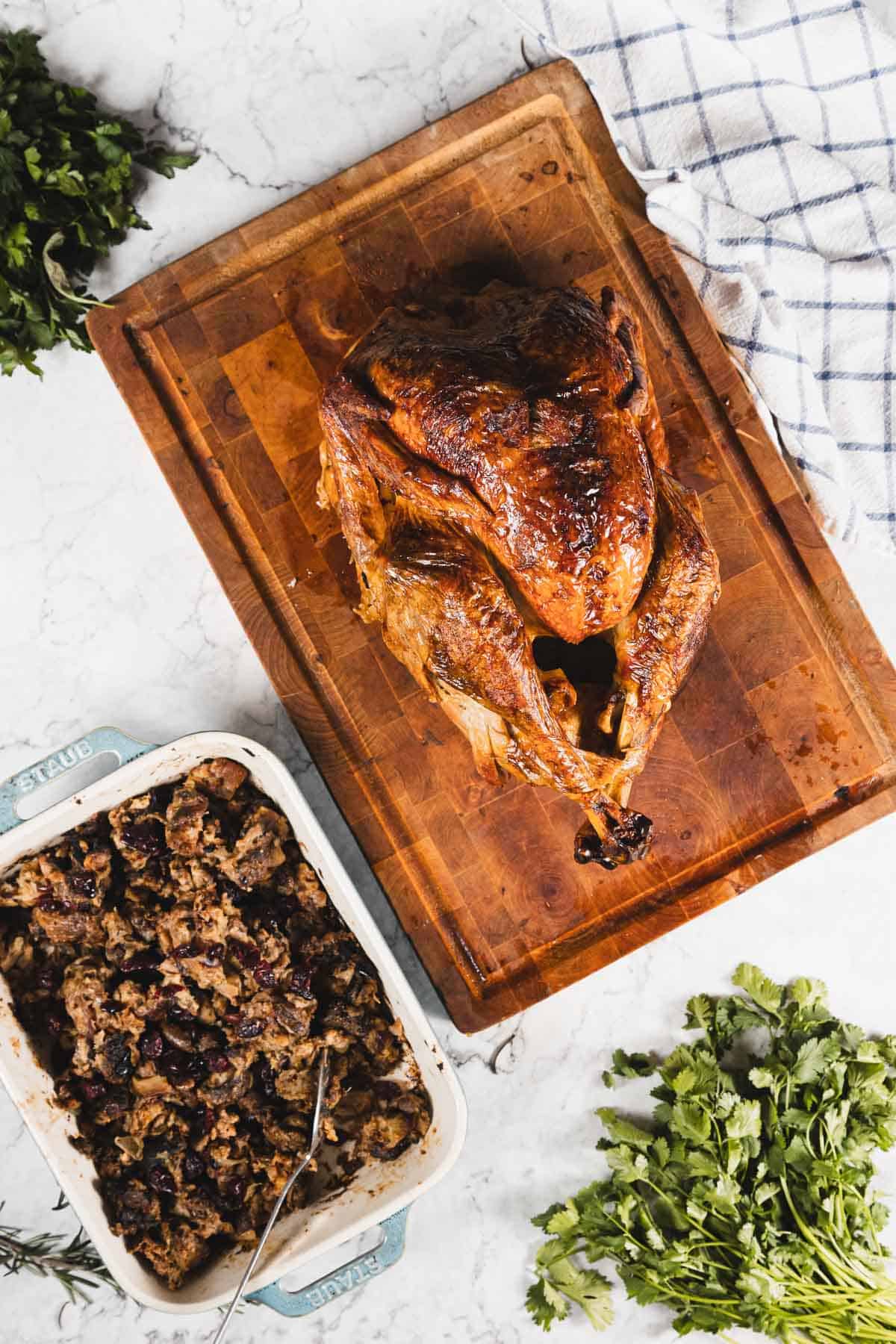 A roasted whole turkey on a wooden cutting board sits next to a baking dish filled with gluten free stuffing. Fresh herbs and a white kitchen towel are nearby on the marble countertop.