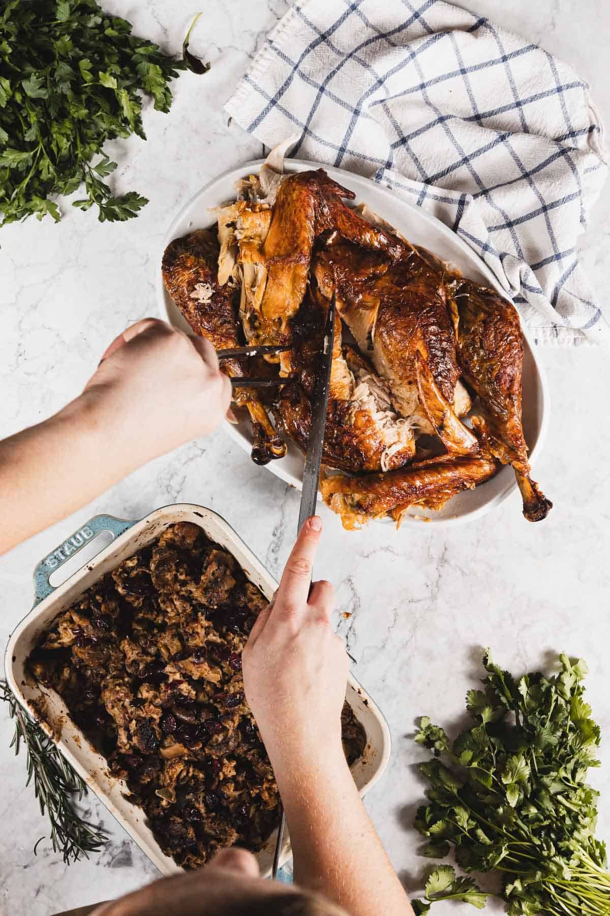 A person uses a knife and fork to carve a roasted turkey on a platter; a baking dish of gluten free stuffing, fresh parsley, and a checkered towel are on a white countertop nearby.