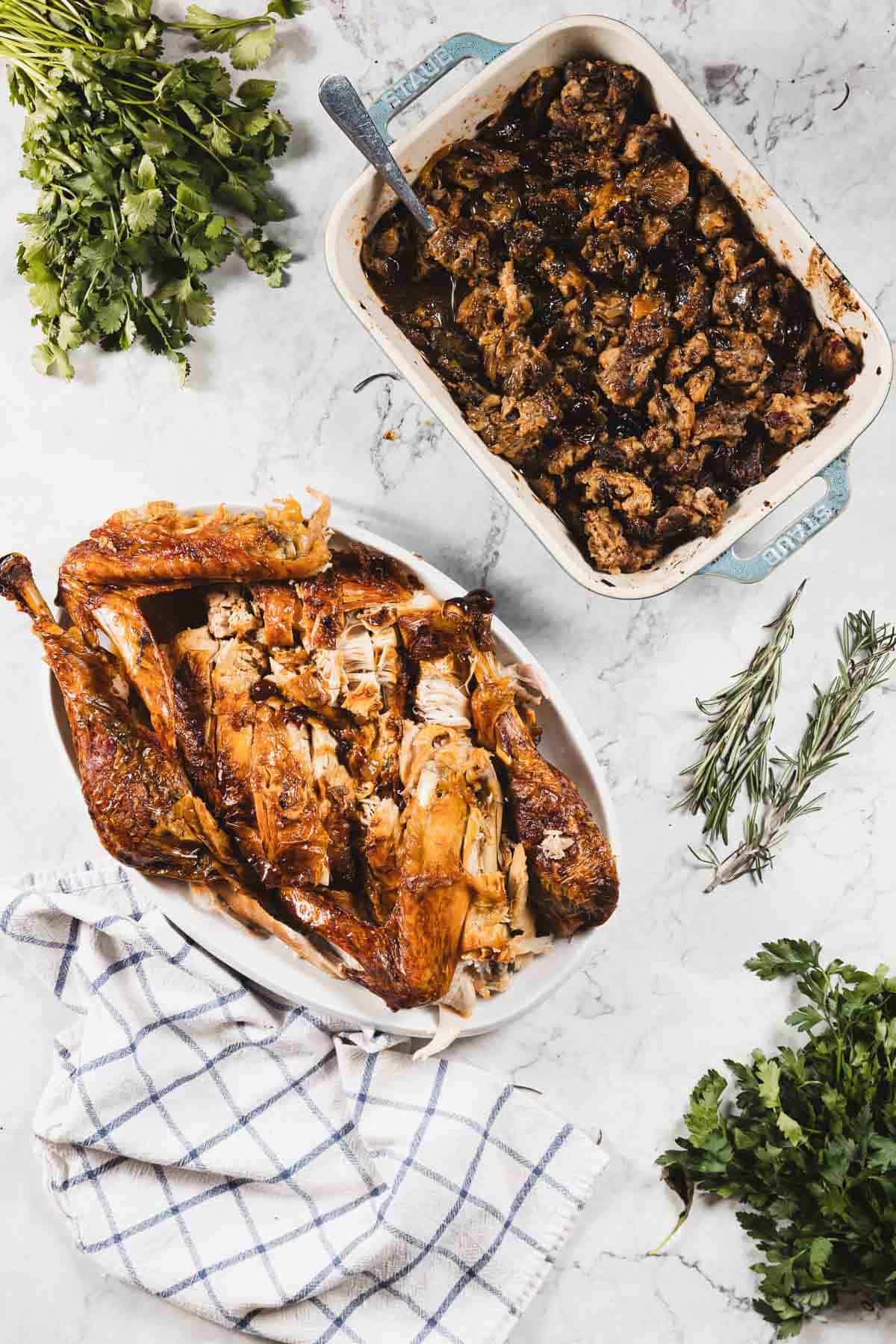 Roast turkey pieces are arranged on a white oval platter next to a baking dish of gluten free stuffing. Fresh herbs, including parsley and rosemary, are scattered on a marble countertop with a checkered towel nearby.