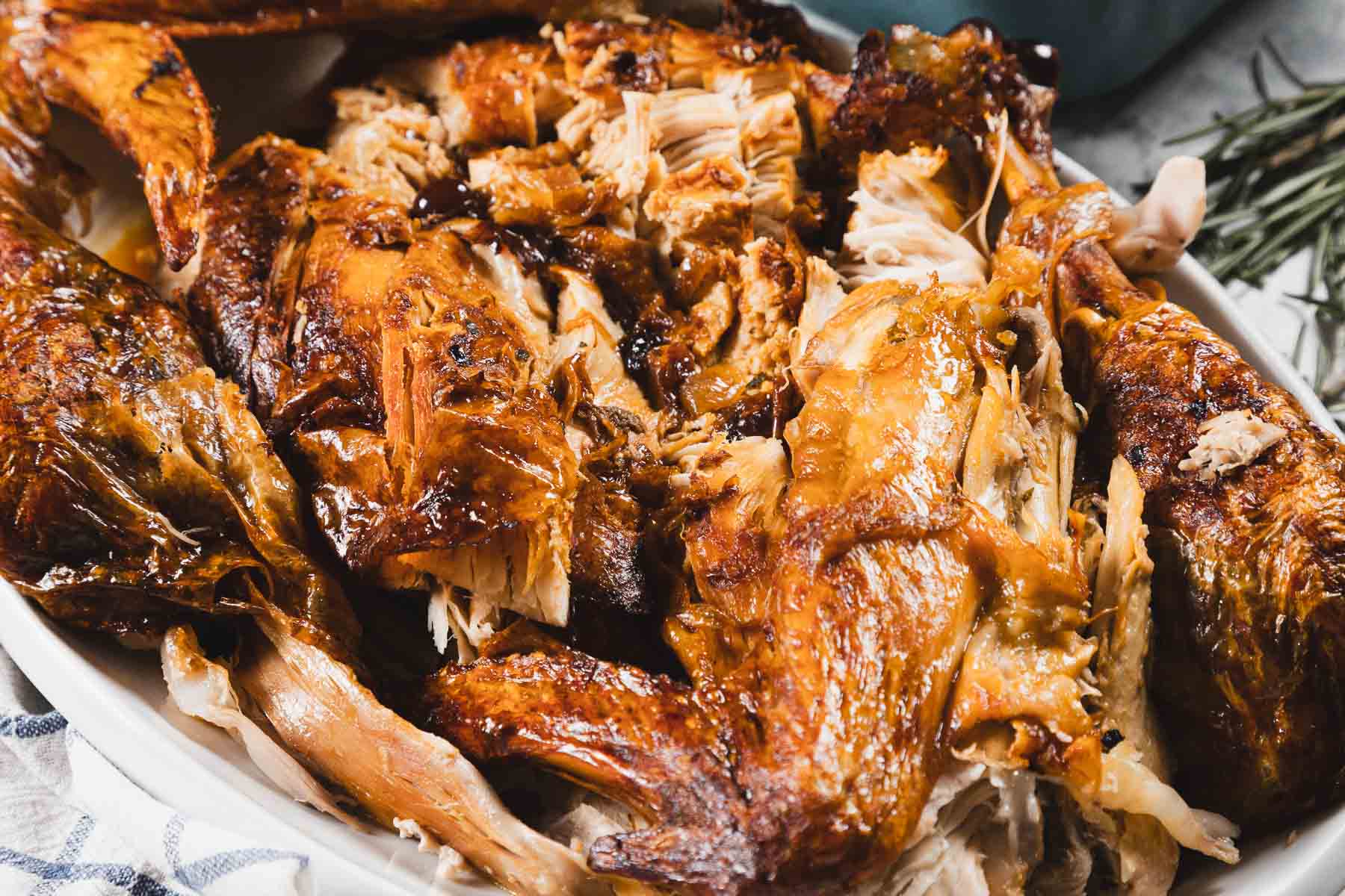 A close-up of a platter filled with pieces of roasted turkey, showing golden-brown, crispy skin and moist, shredded meat. Some rosemary sprigs and gluten free stuffing are visible in the background.