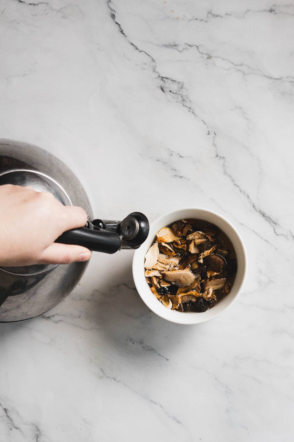 A hand holding a metal kettle is about to pour hot water over dried mushrooms in a white bowl, preparing ingredients for a savory gluten free stuffing on a marble countertop.