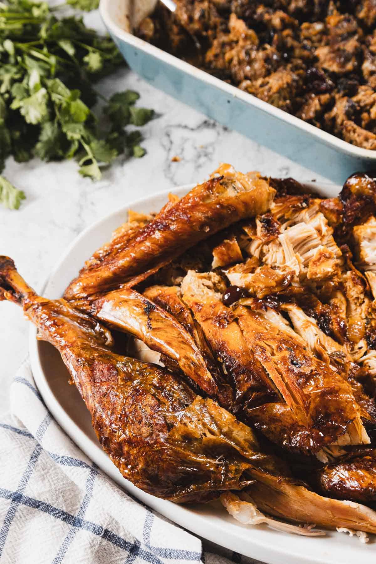 A platter of carved roasted turkey with golden-brown skin&mdash;roasted to perfection with cheesecloth&mdash;sits on a white dish next to a blue towel, with a pan of stuffing and fresh parsley in the background.