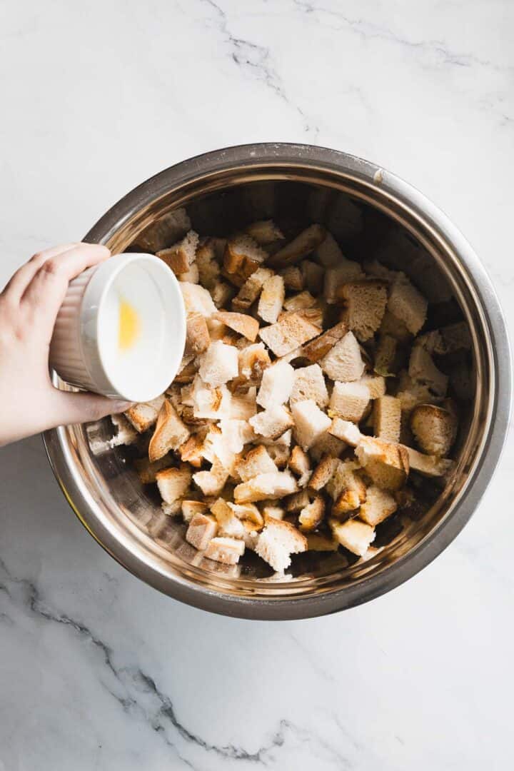 A hand holds a small white bowl with liquid above a large metal bowl filled with cubed pieces of bread for gluten free stuffing on a marble countertop.