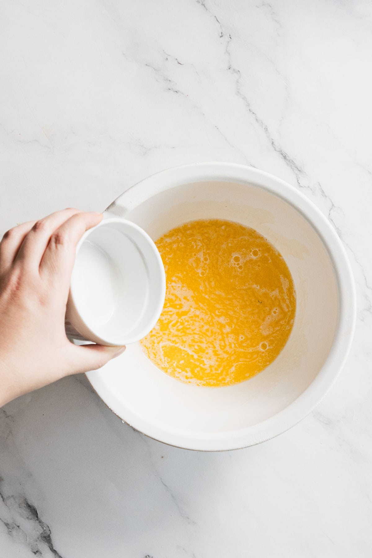 A hand holds a small white bowl, pouring its contents&mdash;often used when preparing turkey or straining with cheesecloth&mdash;into a larger white bowl filled with a yellow liquid mixture on a white marble surface.