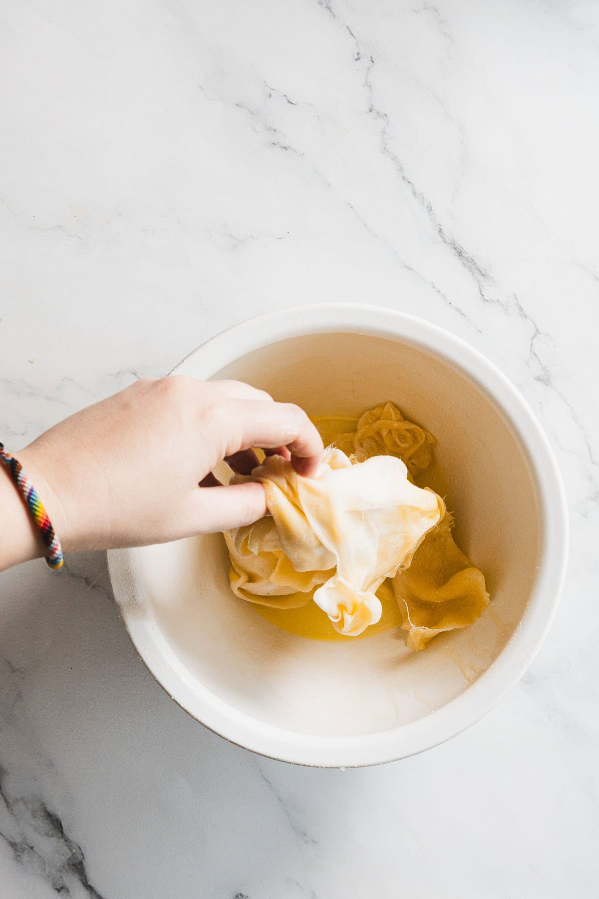 A hand holding a kombucha SCOBY above a white bowl filled with liquid, on a white marble surface&mdash;ideal for straining like turkey through cheesecloth.
