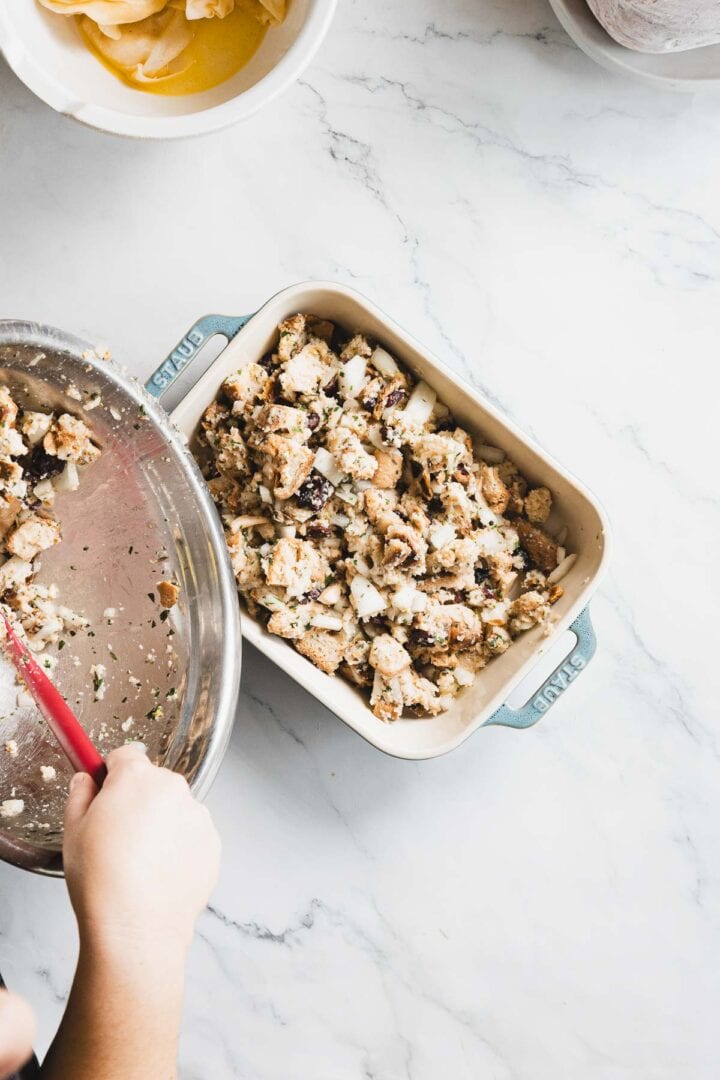 A person uses a red spatula to transfer gluten free stuffing mixture from a metal bowl into a rectangular baking dish on a white marble countertop. Another bowl with sliced ingredients is partially visible above.