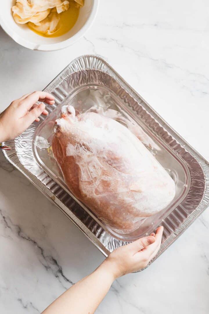 A person holds a large aluminum tray with a frozen whole turkey, ready for the oven. In the upper left corner on the marble surface, a bowl with yellowish gluten free stuffing and a cloth are visible.