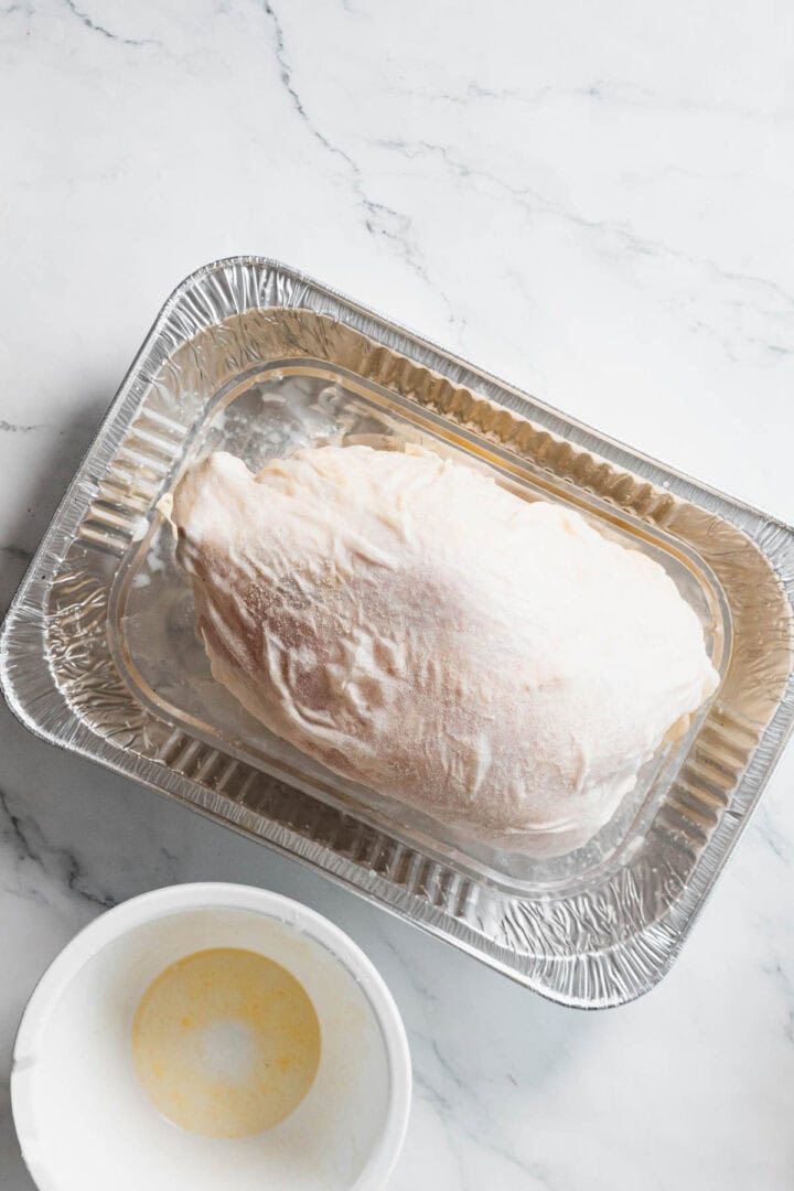 A raw turkey breast covered in a white coating sits in a disposable aluminum roasting pan, ready for the oven. Nearby, a bowl of melted butter awaits alongside gluten-free stuffing on the marble countertop.