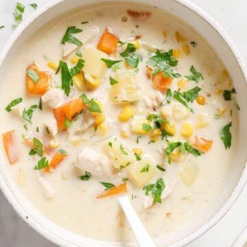 A bowl of creamy soup with visible chunks of carrots, corn, potatoes, chicken, and herbs, with a spoon inside the bowl and chopped parsley sprinkled on top.