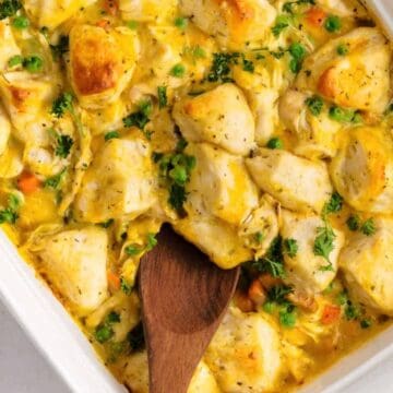 A white baking dish filled with a creamy casserole containing chunks of biscuit, chicken, peas, carrots, and garnished with fresh parsley. A wooden spoon rests in the dish, ready for serving.