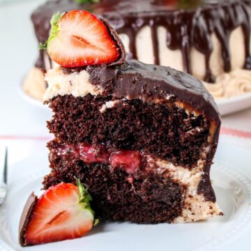 A slice of chocolate cake with chocolate frosting, a layer of strawberry filling, and a halved strawberry on top sits on a white plate. A fork is next to the plate and the whole cake is in the background.