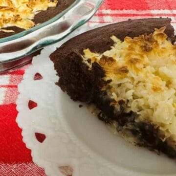 A slice of chocolate and coconut pie sits on a white plate, with the rest in a glass dish nearby. Both are on a red and white checkered cloth—a delicious nod to Boomer recipes with its dark base and toasted coconut topping.