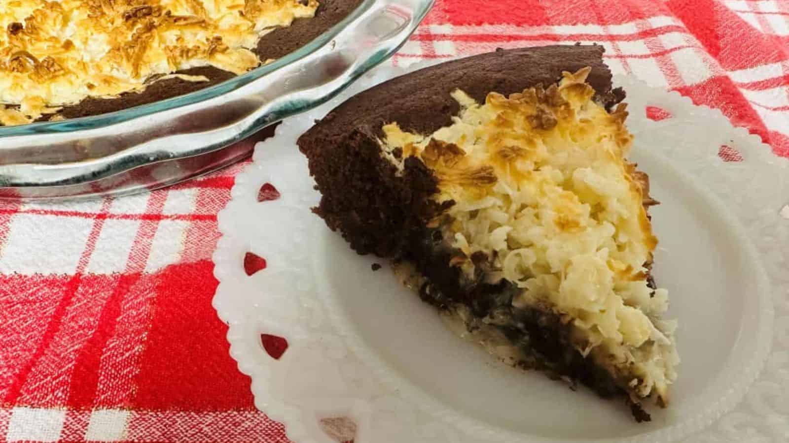 A slice of chocolate and coconut pie sits on a white plate, with the rest in a glass dish nearby. Both are on a red and white checkered cloth—a delicious nod to Boomer recipes with its dark base and toasted coconut topping.