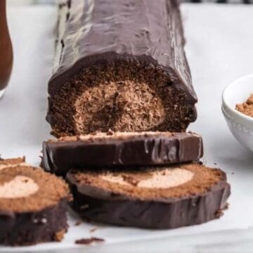 A chocolate Swiss roll cake with chocolate coating is placed on a white surface. Two slices are cut from the roll, showing a swirled chocolate filling. A bowl of cocoa powder and a glass of chocolate drink are nearby.