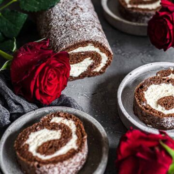A chocolate Swiss roll with cream filling, reminiscent of nostalgic candy enjoyed by Boomers, is placed on a grey surface surrounded by red roses; two slices are served in small grey bowls nearby.
