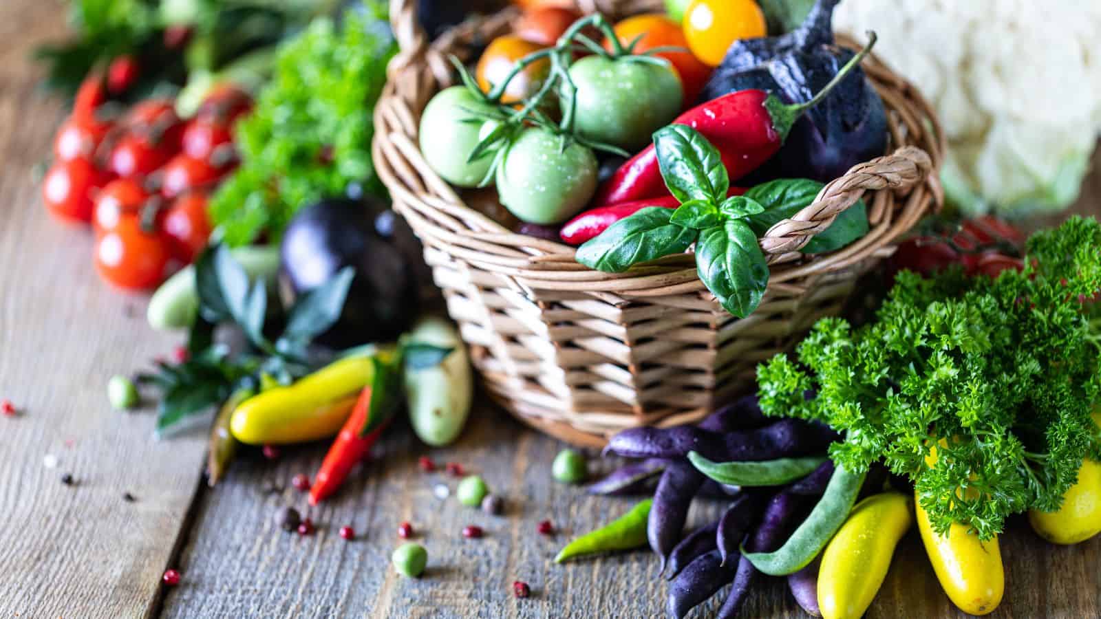 A wicker basket filled with assorted fresh vegetables, including tomatoes, peppers, and basil, sits on a wooden surface surrounded by more vegetables and leafy greens.