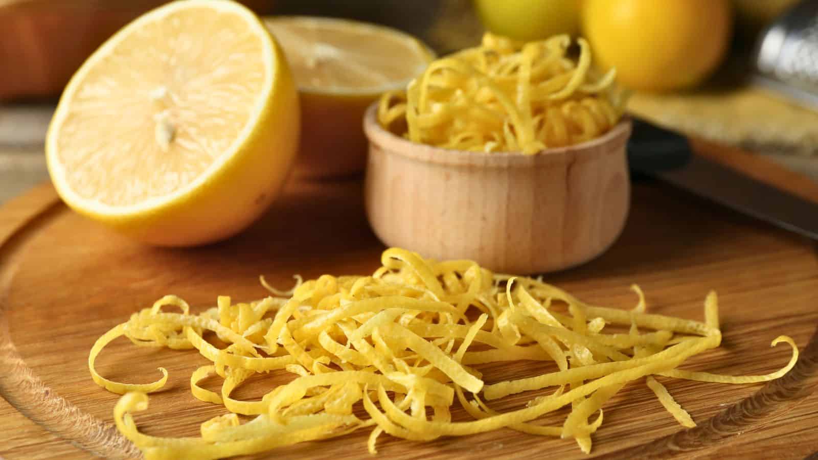 A sliced lemon and a wooden bowl filled with lemon zest strips sit on a wooden cutting board. Loose lemon zest is scattered on the board in front of the bowl. A knife and more lemons are in the background.