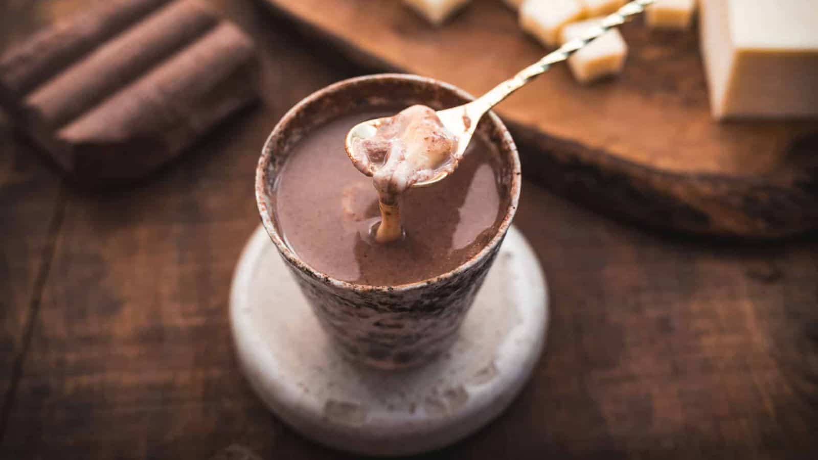 A ceramic cup filled with hot chocolate sits on a round stone coaster. A spoon with melted chocolate is held above the cup. A wooden board with chocolate pieces is in the background.
