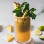 A tall glass filled with Colombian Lulo Juice, garnished with a pineapple wedge, lime slice, and a sprig of fresh mint. Sliced limes and pineapple pieces are scattered in the background.