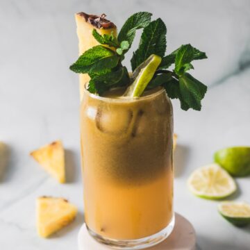 A tall glass filled with Colombian Lulo Juice, garnished with a pineapple wedge, lime slice, and a sprig of fresh mint. Sliced limes and pineapple pieces are scattered in the background.