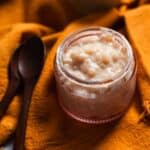 A small glass jar filled with creamy Colombian rice pudding sits on an orange cloth next to two dark wooden spoons.