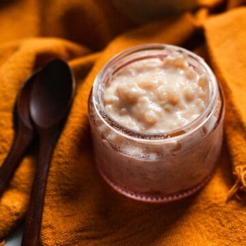 A small glass jar filled with creamy Colombian rice pudding sits on an orange cloth next to two dark wooden spoons.