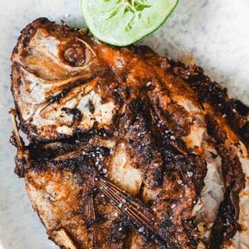 Close-up of a cooked, seasoned Whole Fried Mojarra Frita with crispy skin, garnished with a slice of lime on top, served Colombian style on a light-colored plate.