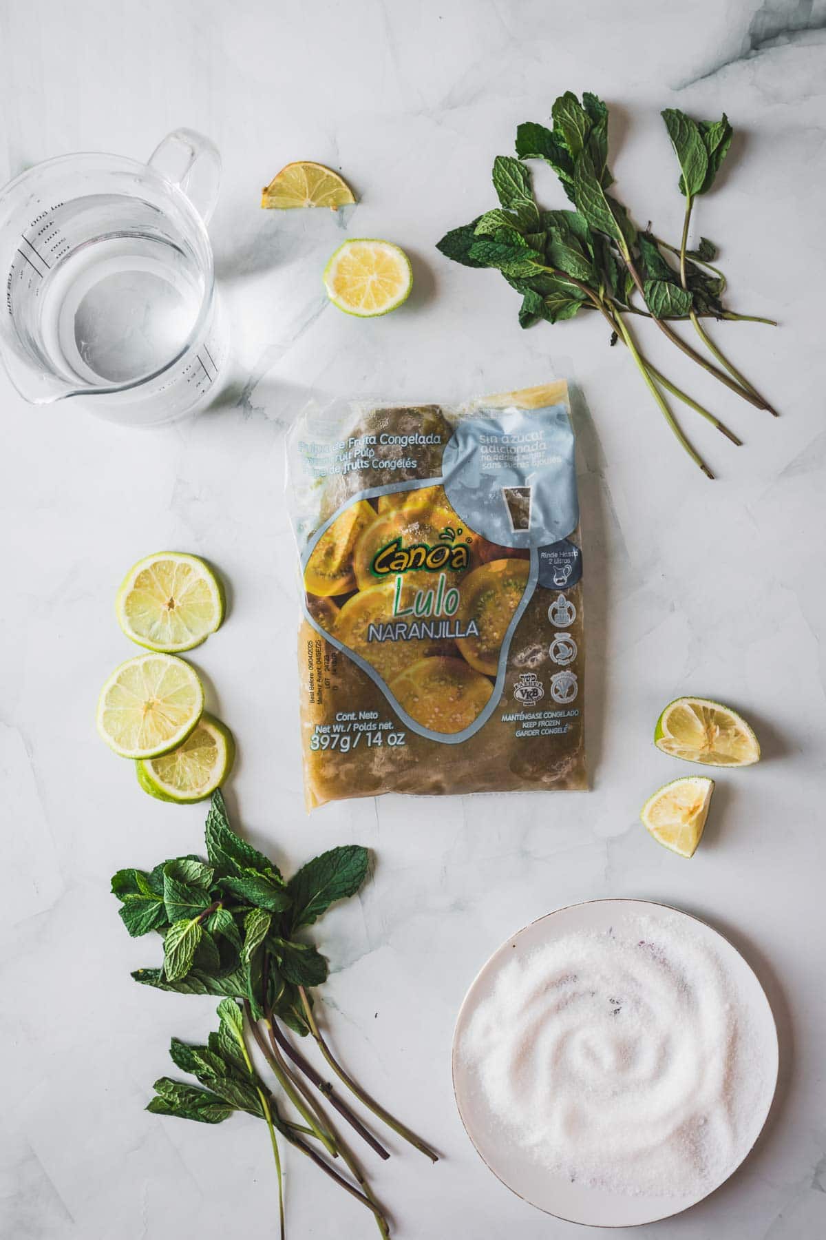 A package of frozen lulo pulp on a marble surface surrounded by fresh mint sprigs, lime slices, a bowl of sugar, and a glass measuring cup filled with water.