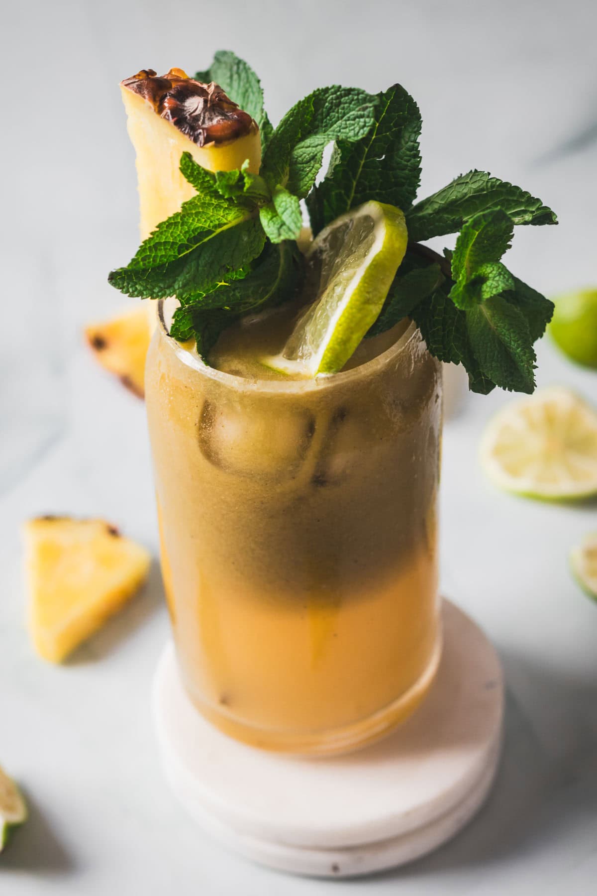 A tall glass filled with an orange-brown drink, garnished with a pineapple wedge, a lemon slice, and a bunch of fresh mint leaves, sits on a white coaster. Pineapple and lime slices are scattered in the background.